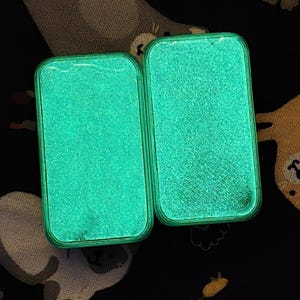 May include: Two open, rectangular cases with rounded corners, each glowing with a bright, speckled, teal-green light. The cases are set against a dark background with a pattern of white and tan shapes.