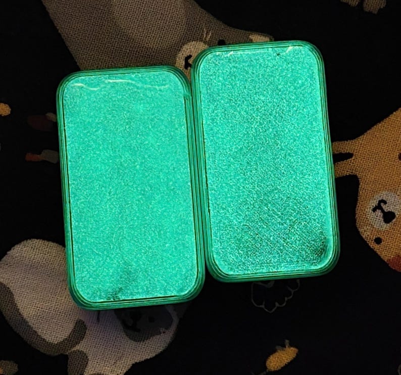 May include: Two open, rectangular cases with rounded corners, each glowing with a bright, speckled, teal-green light. The cases are set against a dark background with a pattern of white and tan shapes.