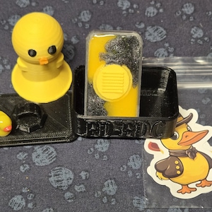 May include: A black and yellow resin mold with a yellow duck figurine and a yellow duck sticker. The mold has the word "JELDOC" printed on it.