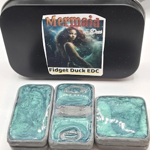 May include: A black tin with a label that reads "Mermaid Dust" and "Fidget Duck EDC" above three small, rectangular fidget toys. The fidget toys have a teal and gray marbled design.