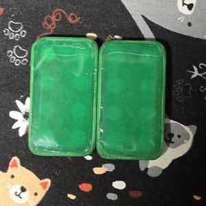 May include: Two rectangular, translucent green cases with rounded corners are displayed. The cases are side-by-side, and the background is a black fabric with a repeating pattern of cartoon cats, paw prints, and flowers.