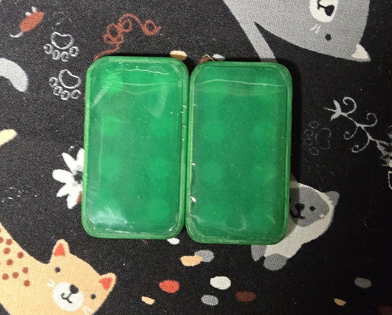 May include: Two rectangular, translucent green cases with rounded corners are displayed. The cases are side-by-side, and the background is a black fabric with a repeating pattern of cartoon cats, paw prints, and flowers.