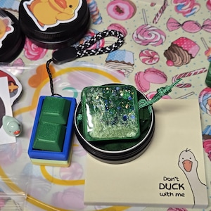 May include: A collection of handmade accessories, including a green and blue square pendant with glitter, a green block-shaped item, and a black tin with a duck sticker. A notepad with a goose illustration and the text "Don't DUCK with me" is also present.