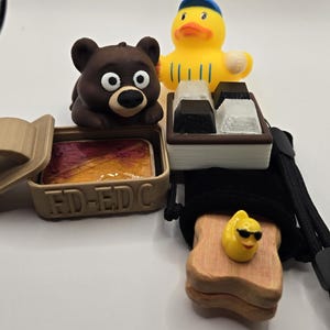 May include: A selection of novelty items, including a brown bear figurine, a yellow rubber duck, and a small wooden box with the text "FD-EDC". The items are arranged on a white surface.