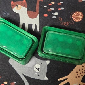 May include: Two green rectangular soap dishes with a transparent, green soap bar inside. The dishes are made of a 3D-printed material. The background is a dark fabric with cat and yarn illustrations.