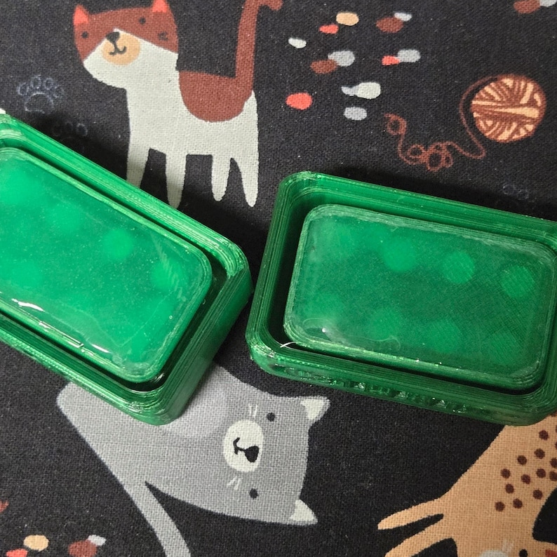 May include: Two green rectangular soap dishes with a transparent, green soap bar inside. The dishes are made of a 3D-printed material. The background is a dark fabric with cat and yarn illustrations.