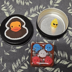 May include: A black tin featuring a duck illustration, a small tin containing a yellow avocado wearing sunglasses, and a small square with red, white, and blue buttons. The items are on a grey patterned surface.