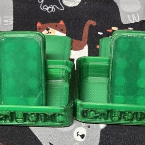 May include: Two green plastic game holders, each with a rectangular piece inside. The holders have the word "CALEDO" printed on the front. The background is a patterned fabric with cartoon animals.
