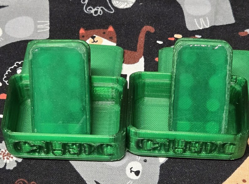 May include: Two green plastic game holders, each with a rectangular piece inside. The holders have the word "CALEDO" printed on the front. The background is a patterned fabric with cartoon animals.