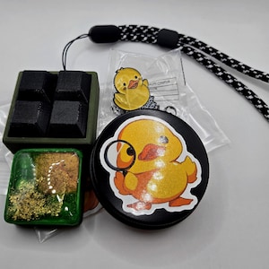 May include: An assortment of items including a black and white lanyard, a green and gold resin square, a black container with four black squares, a yellow duck pin, and a black pop-up phone grip with a yellow duck illustration.