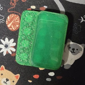 May include: Two green rectangular phone cases. One case has a textured, glittery surface, while the other is smooth. The cases are placed on a black fabric with a pattern of white flowers, yarn balls, and cartoon animals.