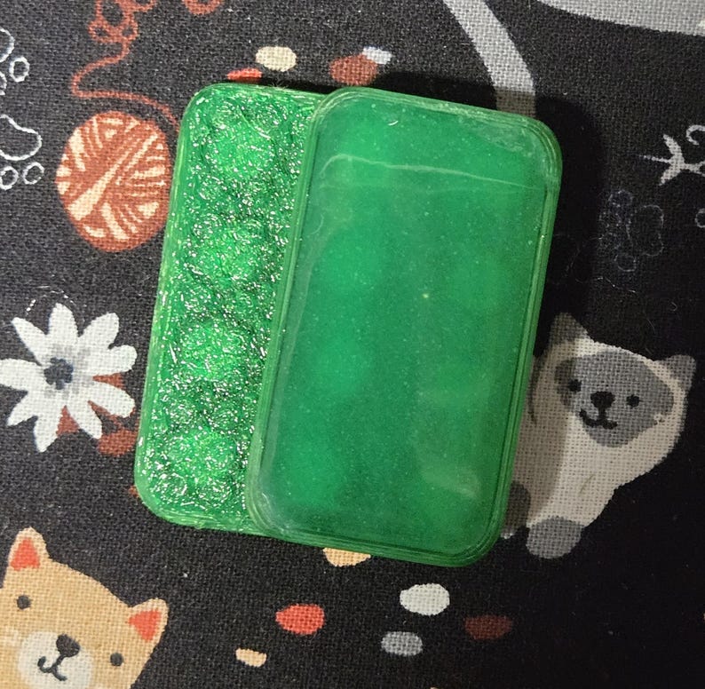 May include: Two green rectangular phone cases. One case has a textured, glittery surface, while the other is smooth. The cases are placed on a black fabric with a pattern of white flowers, yarn balls, and cartoon animals.