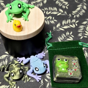 以下が含まれることがあります： 黒い円筒形の容器で、明るい木製の蓋の上には緑色のカエルと黄色いアヒルが乗っています。その横には、メタリックグリーンのカエル、水色のカエル、そして緑色のベルベットの袋の上に置かれた4匹のカエルのフィギュアが入った小さな正方形の箱があります。