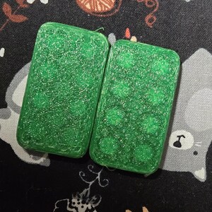 May include: Two rectangular, green, glittery resin dominoes. One domino has a smooth surface, while the other has a pattern of green dots. The dominoes are set against a dark background with a floral and cat pattern.