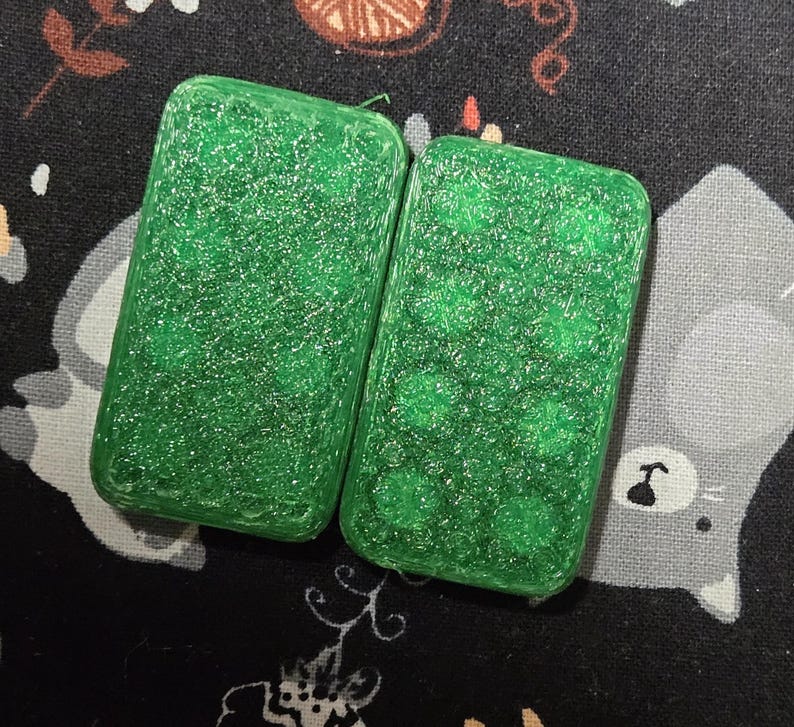 May include: Two rectangular, green, glittery resin dominoes. One domino has a smooth surface, while the other has a pattern of green dots. The dominoes are set against a dark background with a floral and cat pattern.