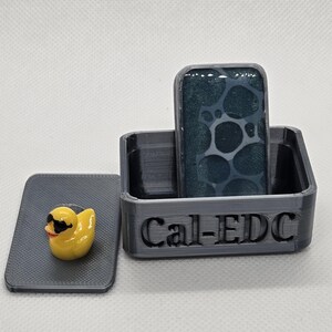 May include: A dark gray box with the text "Cal-EDC" holds a rectangular item with a dark teal pattern. A yellow rubber ducky wearing sunglasses rests on a gray lid.