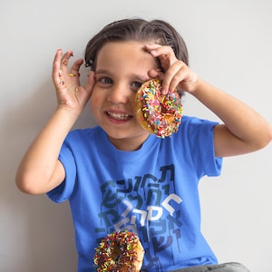May include: A young boy wearing a blue t-shirt with a graphic print of black and white letters. He is holding a donut with colorful sprinkles in front of his face and smiling.