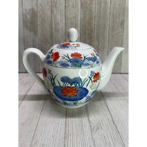Può includere: Teiera in ceramica bianca con coperchio e manico. La teiera è decorata con un motivo floreale con fiori rossi, blu e arancioni e foglie verdi. La teiera è appoggiata su una superficie di legno.