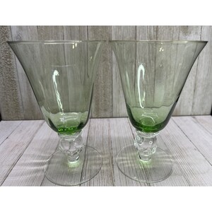 May include: Two matching goblets made of light green glass. Each goblet features a wide, flared bowl with vertical ribbing, a clear glass stem with a decorative knop detail, and a circular clear glass base for stability on flat surfaces.