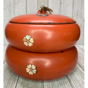 Puede incluir: Un recipiente redondo de color naranja de dos niveles con una tapa que tiene una pequeña borla marrón. Cada nivel está decorado con una flor incrustada de cuatro pétalos hecha de nácar iridiscente. La tapa también muestra un pequeño motivo floral circular.