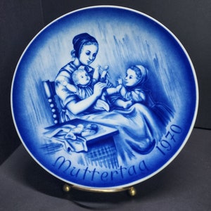 May include: Blue and white porcelain plate with a mother and two children. The mother is holding a baby and the other child is sitting on her lap. The plate has the text "Muttertag 1970" on the bottom.