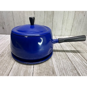 May include: A cobalt blue enamel saucepan with a lid and a black handle. The saucepan sits on a matching blue base. The handle is black and angled. The lid has a black knob.