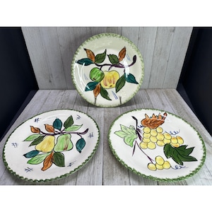 以下が含まれることがあります： 果物のデザインが施された3つのヴィンテージセラミックプレート。プレートには、リンゴ、梨、ブドウの手描きのイラストが、緑と茶色の葉とともに描かれています。各プレートには緑色のスカラップエッジがあります。プレートは木製の表面にあります。