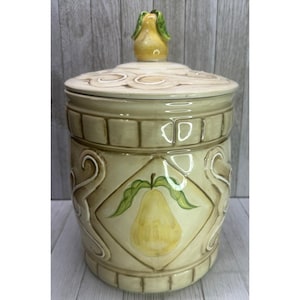May include: A ceramic cookie jar with a lid, featuring a pear design. The jar is primarily beige with brown accents and a yellow pear illustration. The lid has a pear-shaped handle and decorative swirls.