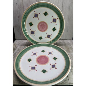 May include: Two white ceramic plates with a green and tan border. Each plate features a pink center circle and is decorated with green and purple diamond shapes. The plates are on a wooden surface.