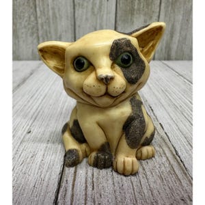 May include: A cream-colored cat figurine with black spots and green eyes. The cat is sitting upright with its ears perked up. The figurine has a smiling expression and is made of a smooth material.