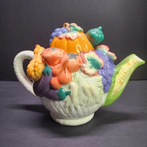 May include: A ceramic teapot with a white handle and spout, adorned with colorful, three-dimensional vegetables. The teapot features a pumpkin, tomatoes, corn, eggplant, and grapes, creating a whimsical, harvest-themed design.