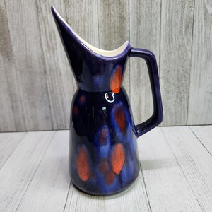May include: A ceramic pitcher with a dark blue glaze and orange accents. The pitcher has a unique, elongated spout and a handle. The interior is white. The pitcher is likely for serving beverages.