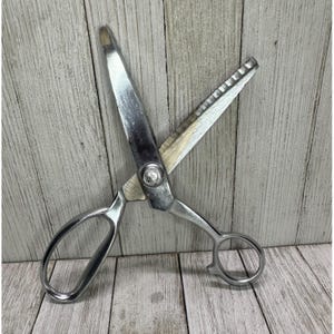 May include: A pair of large, silver-colored scissors with a rounded handle design. The blades are sharp and open, with one blade featuring a serrated edge. The scissors are designed for cutting fabric or other materials.
