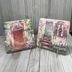 May include: Two decorative ceramic plates with painted scenes of Italian-style buildings. One plate features a red door with a window above it, while the other plate shows a blue door with a window and steps leading up to it. Both plates have a floral design around the edges.