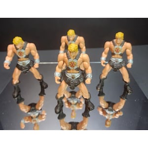 May include: Multiple miniature action figures of a muscular, blonde-haired character in a brown and black outfit. The figures are standing on a reflective surface, with their reflections visible. The figures have a shield-like emblem on their chest.