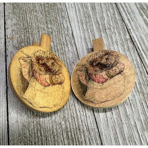 May include: Two circular wooden clips featuring a pyrography illustration of a hedgehog tucked into a fabric pouch. The hedgehog has detailed brown quills and a reddish-patterned wrap, while the wooden base has a natural light tan finish.