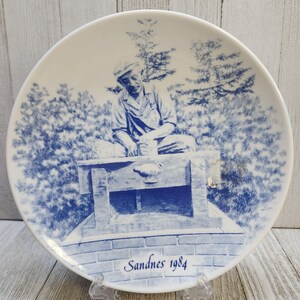 May include: A blue and white ceramic plate with a depiction of a man sitting on a stone bench. The plate has the text "Sandnes 1984" on the bottom.