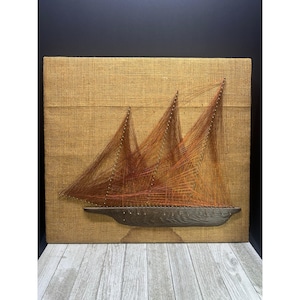 May include: A string art sailboat with three sails, crafted with orange thread on a tan burlap canvas. The boat's hull is dark brown. The artwork is framed in a dark colour. The piece is a unique, handmade nautical decoration.