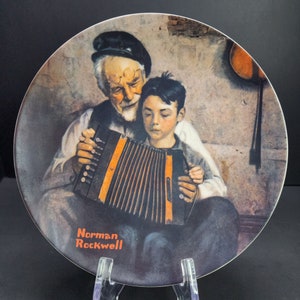 Puede incluir: Un plato coleccionable de estilo vintage con una pintura de Norman Rockwell de un hombre mayor y un niño pequeño tocando un acordeón. El plato tiene un fondo marrón con una pared marrón claro y un suelo marrón oscuro. El hombre lleva un sombrero azul oscuro y una chaqueta marrón. El niño lleva una camisa azul claro y una chaqueta azul oscuro. El acordeón es marrón y tiene un diseño de acordeón blanco y negro. El plato tiene las palabras "Norman Rockwell" impresas en naranja en la parte inferior.