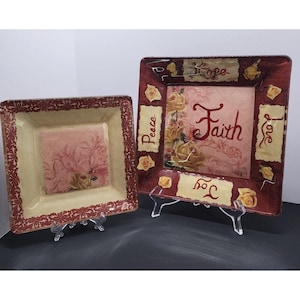 Può includere: Due piatti decorativi quadrati con un bordo rosso scuro a motivo floreale. Il piatto più grande mostra le parole Faith, Hope, Peace, Love e Joy accanto a illustrazioni di rose gialle. Il piatto più piccolo presenta un centro floreale rosa.