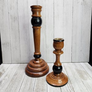 May include: Two wooden candle holders. The taller one has a long, slender body with a bulbous section painted black. The shorter candle holder has a similar design. Both are made of wood and have a warm, brown tone.