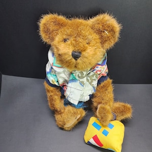Op de afbeelding: Een bruine teddybeer die een blauw-wit gestreept shirt draagt met een geel-blauw gestreept kussen.