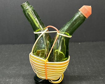 May include: Two green glass bottles with red tops are held together by a woven basket made of orange and white string.
