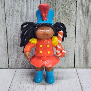 May include: A Cabbage Patch Kids doll in a marching band uniform. The doll has black yarn hair, brown skin, and is wearing a red jacket, blue boots, and a blue hat with a red plume. It holds a red and white striped candy cane.