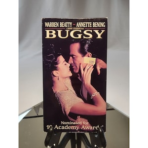 Può includere: Una custodia VHS con una foto in bianco e nero di Warren Beatty e Annette Bening in un abbraccio romantico. Il titolo "BUGSY" è in grandi caratteri grassetto. Il testo "Nominato per 10 premi Oscar" è in fondo alla custodia.