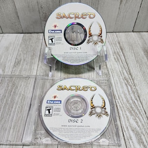 Puede incluir: Dos discos de juego de ordenador plateados para el juego Sacred. Los discos están etiquetados como "Disco 1" y "Disco 2" y presentan el logotipo del juego y un diseño de insecto estilizado.