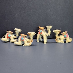 May include: A collection of five small, beige felt camels with brown saddlebags and turquoise accents. The camels are in various poses, some kneeling and others standing. Each camel has a detailed, stitched design.