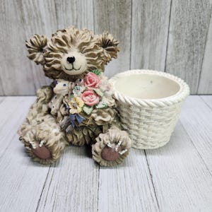May include: A ceramic teddy bear figurine holding a bouquet of flowers, sitting next to a white wicker planter. The planter has a smooth, white finish and a woven texture.