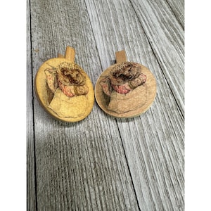 May include: Two wooden ornaments with a brown hedgehog illustration. The hedgehog is wearing a pink and white dress and is holding a cup. The ornaments are round and have a light brown wood grain.