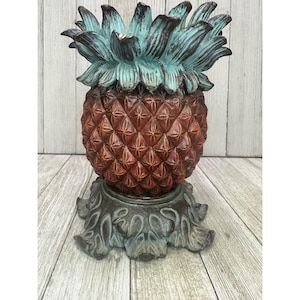 Op de afbeelding: Een decoratieve ananassculptuur met een roodbruin lichaam en turquoise bladeren. De ananas staat op een decoratieve voet met een verweerde afwerking. De sculptuur is gemaakt van een duurzaam materiaal en is geschikt voor weergave.